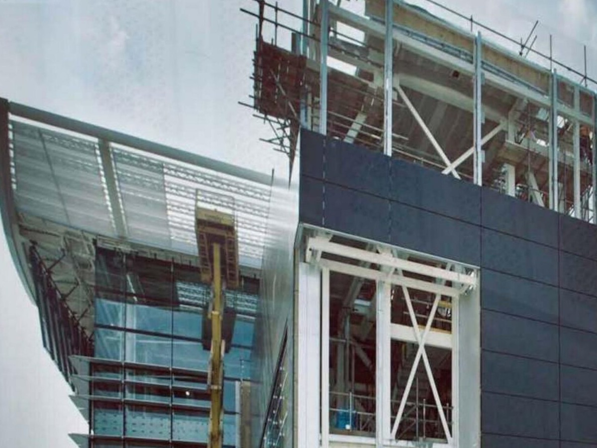 A partially clad building elevation mid-installation, with dark rainscreen and cladding panels fixed alongside exposed structural steel framing and a scaffold access platform.