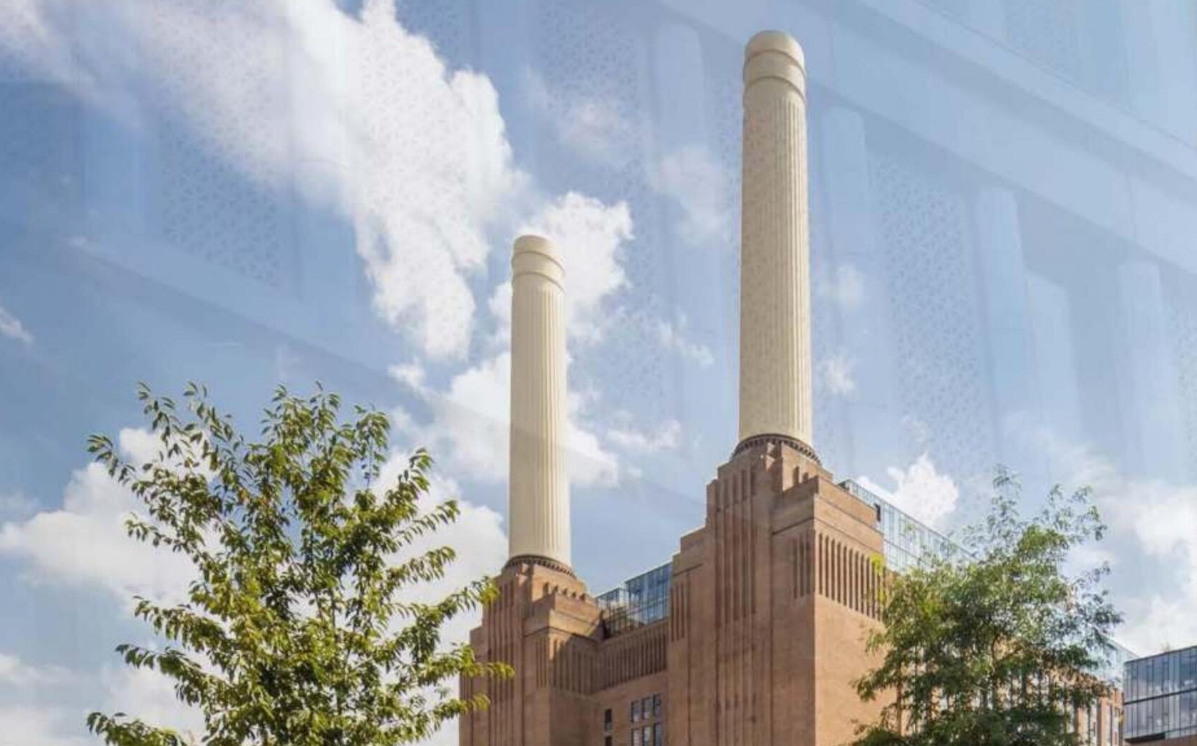 A glazed building facade reflecting a brick industrial-style structure with tall chimneys and modern glazed extensions beyond, trees in the foreground under a partly clouded sky — a facade and cladding installation photograph.